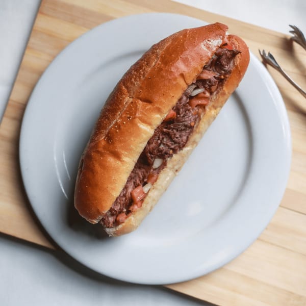 A beef sandwich with tomatoes and onions on a white plate, with tongs on the side.