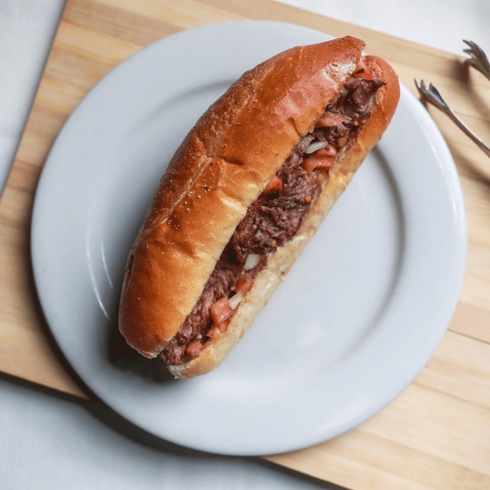 A beef sandwich with tomatoes and onions on a white plate, with tongs on the side.