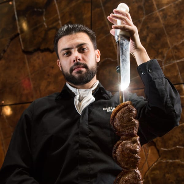 A churrasqueiro holding a skewer of picanha, ready to serve, against a warm, textured background.