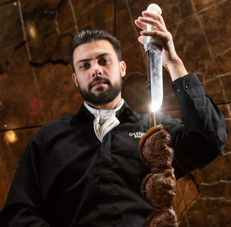 A churrasqueiro holding a skewer of picanha, ready to serve, against a warm, textured background.