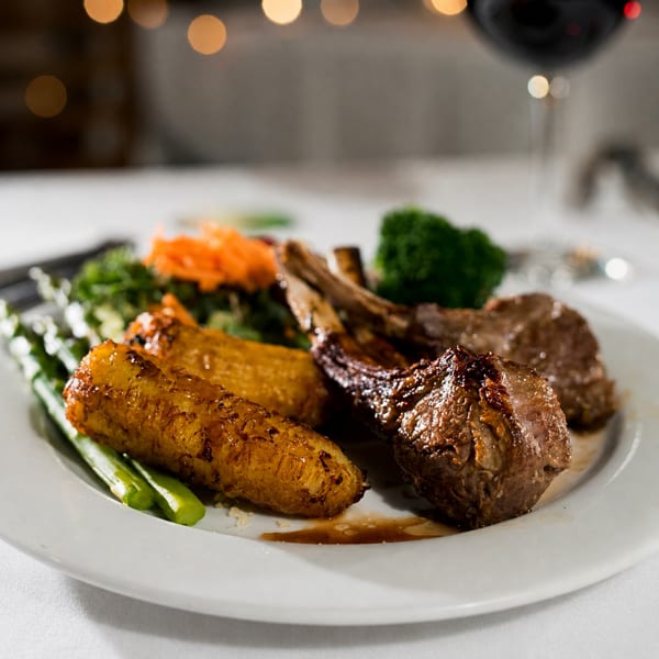 Grilled lamb chops with roasted plantains, asparagus, and broccoli on a white plate.