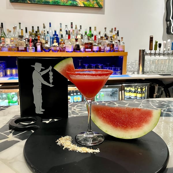 Watermelon martini with a watermelon slice and coconut flakes on a black tray at a bar.