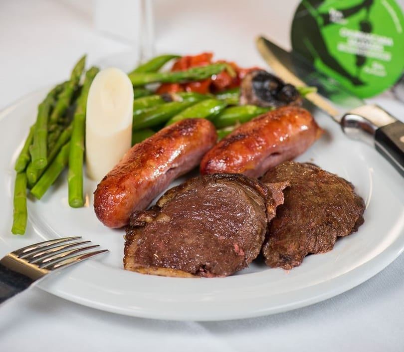 A plate of grilled steak, sausages, asparagus, and heart of palm on a white tablecloth.