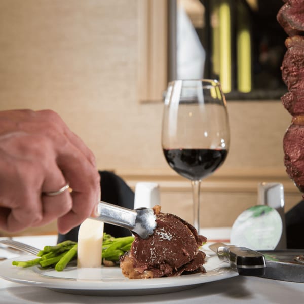 A hand uses tongs to serve sliced steak onto a plate with asparagus and a glass of red wine.