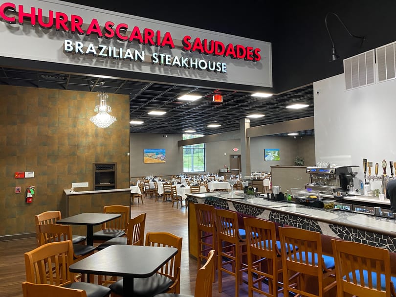 Interior of Churrascaria Saudades Brazilian Steakhouse with dining tables, bar, and chandelier.