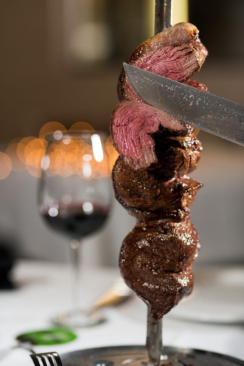 A knife slices a piece of perfectly cooked picanha from a skewer, with a glass of red wine in the background.